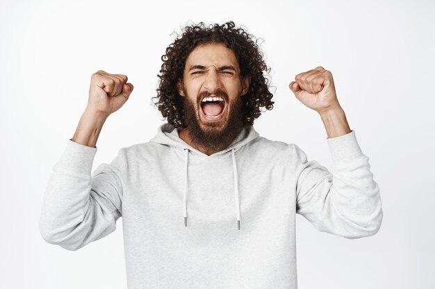 Portret van een vrolijke Spaanse man die schreeuwend van vreugde gebalde vuisten wint en triomfantelijk op een witte achtergrond staat