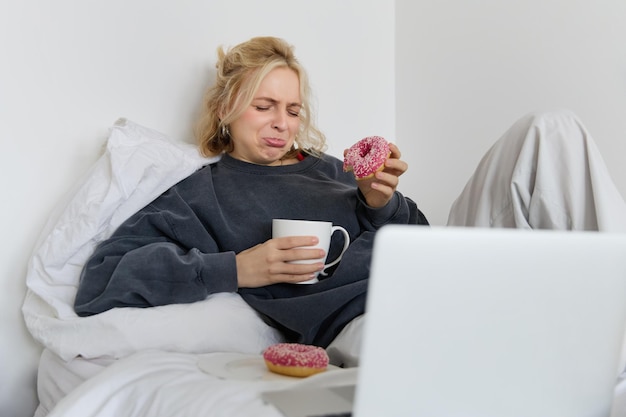 Portret van een vrouw die een droevige film op haar laptop kijkt, huilt en haar tranen afveegt, donuts eet en...