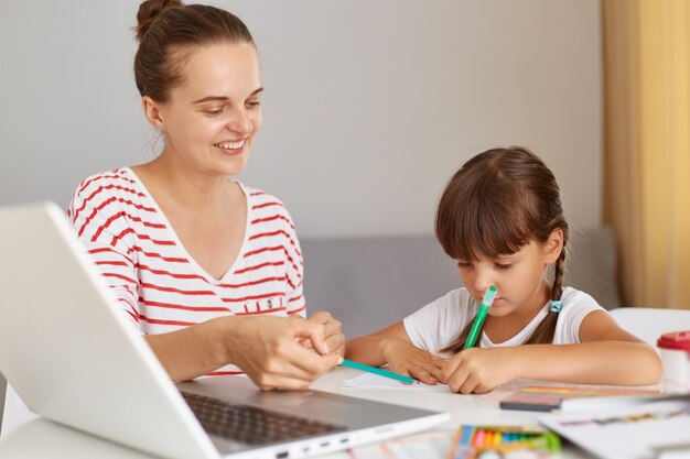 Portret van een vrouw die gestreepte vrijetijdskleding draagt die huiswerk maakt met haar charmante kind, schoolmeisje die thuistaak schrijft in oefeningen, mensen poseren in lichte kamer thuis, online lessen op afstand.