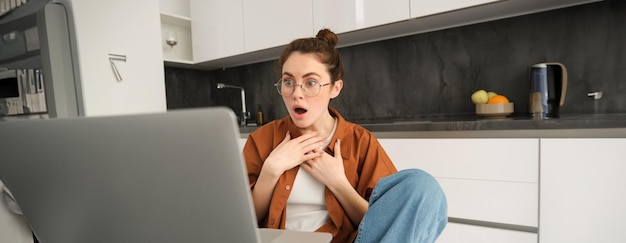 Gratis foto portret van een vrouw die naar een laptop kijkt met een geschokt gezicht en groot nieuws ziet van een vrouwelijke student in de keuken