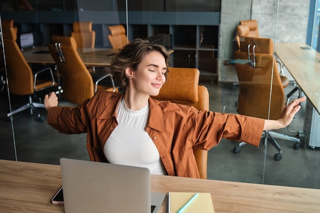 Gratis foto portret van een vrouw die op kantoor rust en haar armen uitstrekt na het zitten en werken op een laptop