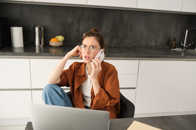 Gratis foto portret van een vrouw die vanuit huis werkt in een bril en met een verward gezicht zit na het beantwoorden van een telefoon