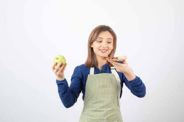 Portret van een vrouw in een schort die probeert te kiezen wat ze appel of pizza gaat eten