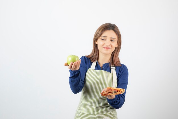 Portret van een vrouw in een schort die probeert te kiezen wat ze appel of pizza gaat eten