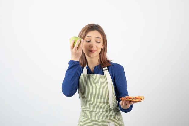 Portret van een vrouw in een schort die probeert te kiezen wat ze appel of pizza gaat eten