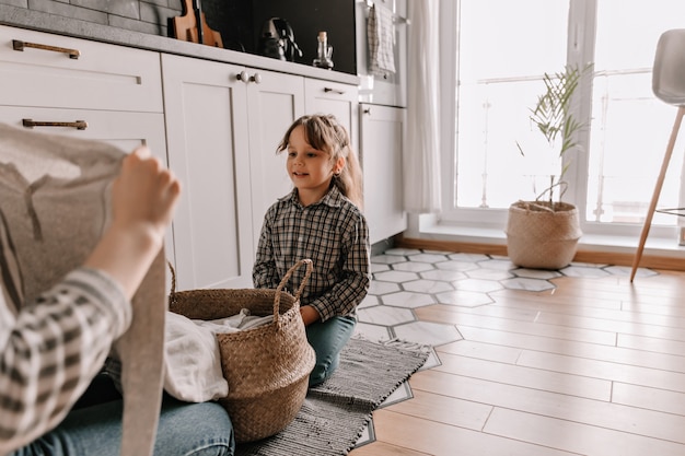 Portret van een vrouw in geruite overhemd zittend op de vloer van de keuken en kijken naar haar moeder kleren uit de mand halen.