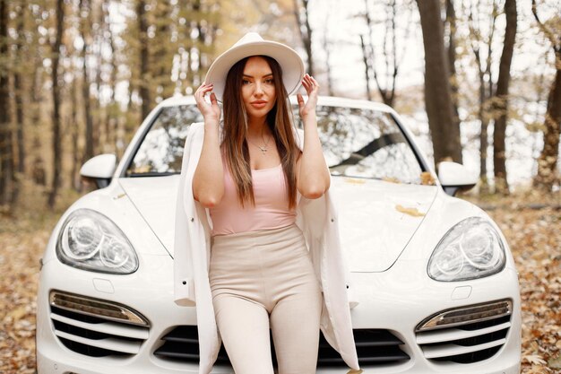 Portret van een vrouw in het herfstbos, die voor haar witte auto staat. Brunette vrouw poseren voor een foto. Vrouw met witte hoed, roze t-shirt en broek.