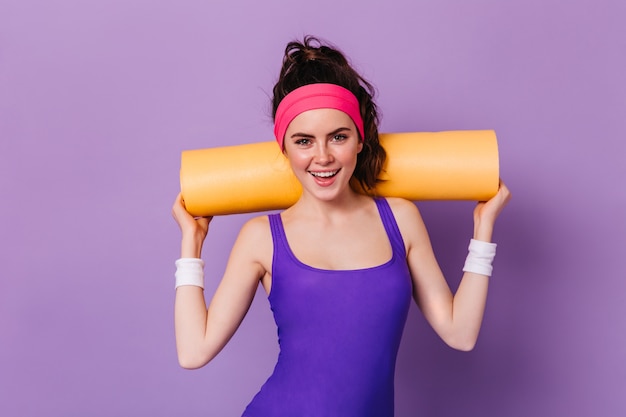 Portret van een vrouw in roze hoofdband voor sport en paarse top, poseren met yoga mat op paarse muur