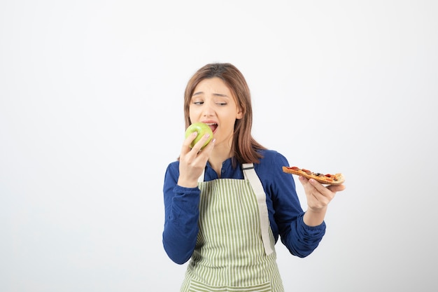 Portret van een vrouw in schort die groene appel eet terwijl ze naar pizza kijkt