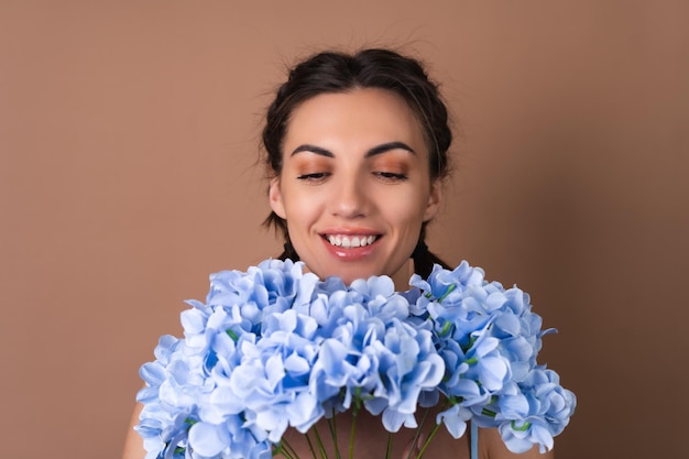 Portret van een vrouw met een perfecte huid en natuurlijke make-up op een beige achtergrond met staartjes in een jurk met een boeket blauwe bloemen