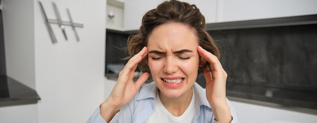 Gratis foto portret van een vrouw met hoofdpijn die in de keuken zit, haar hoofd aanraakt en grimassen maakt, pijnlijke migraine heeft