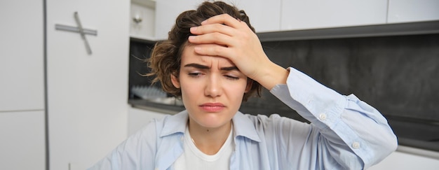 Gratis foto portret van een vrouw met hoofdpijn die in de keuken zit, haar hoofd aanraakt en grimassen maakt, pijnlijke migraine heeft