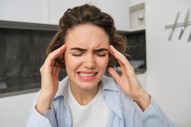 Gratis foto portret van een vrouw met hoofdpijn zit in de keuken, raakt het hoofd aan en grimassen heeft pijnlijke migraine