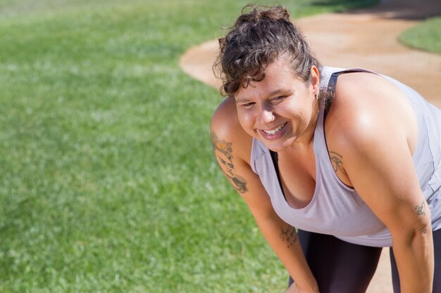 Portret van een vrouw met krullend haar die sport doet op een zonnige dag. Mollige vrouw in grijze top met tatoeages die zijwaarts kijken. Sport, lichaam positief concept