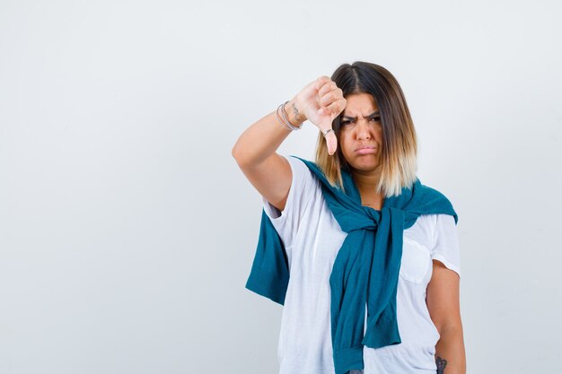 Portret van een vrouw met vastgebonden trui met duim naar beneden, gebogen onderlip in wit t-shirt en teleurgesteld vooraanzicht