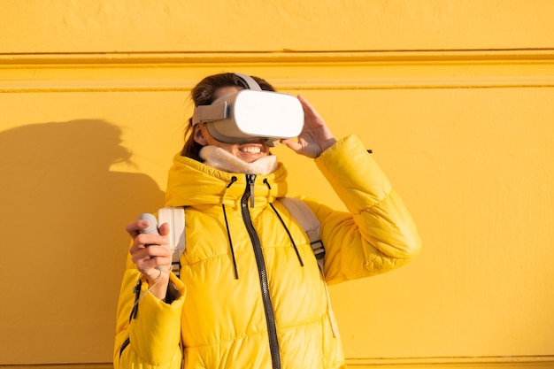 Portret van een vrouw met vr-bril op straat tegen een gele muur in fel zonlicht in de winter in warme kleren