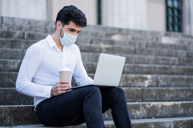 Portret van een zakenman die gezichtsmasker draagt en zijn laptop gebruikt terwijl hij buiten op de trap zit. Bedrijfsconcept. Nieuw normaal levensstijlconcept.