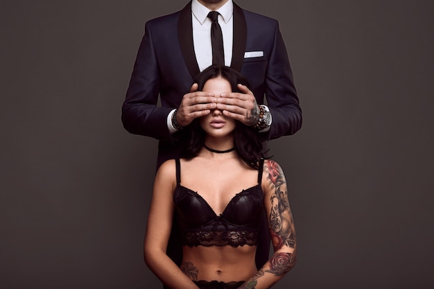 Portret van een zakenman in elegant pak bedek de ogen van sexy vrouw met een tatoeage in lingerie