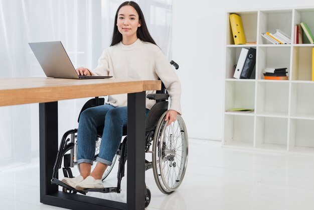 Portret van een zekere jonge onderneemsterzitting op rolstoel die laptop in het bureau met behulp van