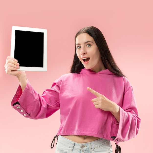 Portret van een zelfverzekerd toevallig meisje dat het lege scherm van laptop toont dat over roze ruimte wordt geïsoleerd