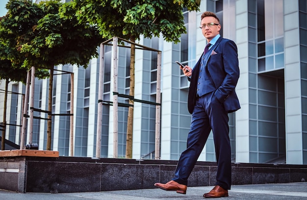 Portret van een zelfverzekerde stijlvolle zakenman gekleed in een elegant pak houdt een smartphone vast en kijkt weg terwijl hij buiten staat tegen de achtergrond van een wolkenkrabber.