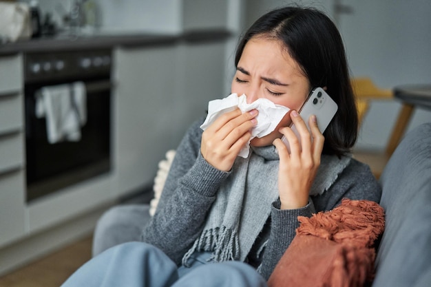Gratis foto portret van een zieke jonge koreaanse vrouw die zich ziek voelt niezen en een servet vasthoudt terwijl ze thuis ziek blijft