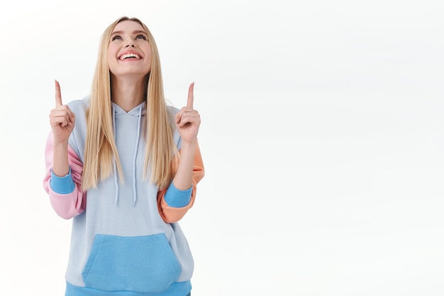 Portret van een zorgeloze, gelukkige knappe blonde vrouw in hoodie, hoofd omhoog, breed glimlachend, met de vingers wijzend en omhoog kijkend