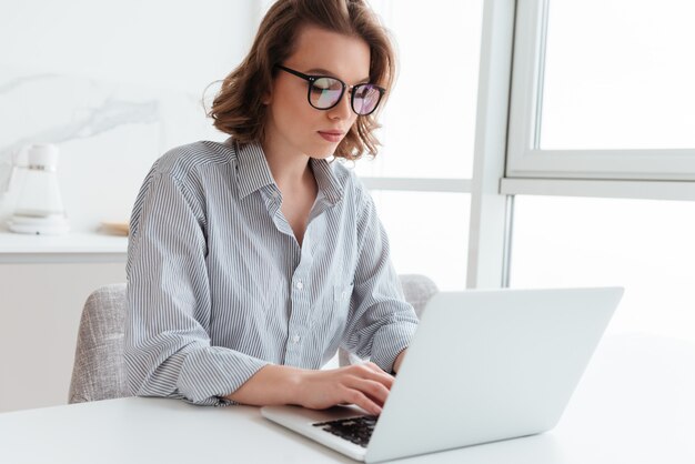 Portret van elegante jonge vrouw die e-mail op laptop texting terwijl het zitten bij lijst in lichte ruimte