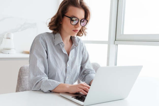 Portret van elegante jonge vrouw die e-mail op laptop texting terwijl het zitten bij lijst in lichte ruimte