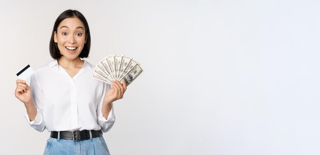 Portret van enthousiaste aziatische vrouw die geld in contanten en creditcard aanhoudt, glimlachend verbaasd over de witte achtergrond van de camera