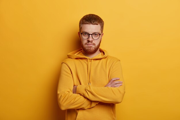 Portret van ernstig uitziende man staat in zelfverzekerd gebaar, houdt de armen over elkaar, heeft een optische bril en een gemberbaard, draagt een hoodie, vormt tegen de gele muur. heeft een assertieve uitdrukking.