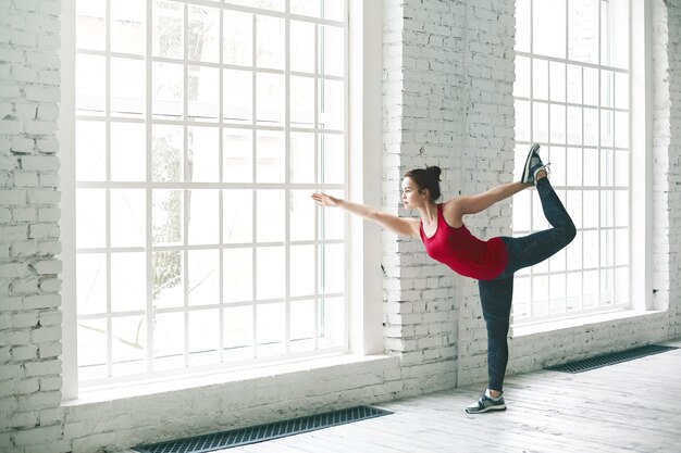 Portret van ernstige geconcentreerde jonge vrouwelijke yogi met mooi prefect atletisch lichaam trainen in het fitnesscentrum, staande in Natarajasana-oefening of Lord of the Dance pose bij groot raam