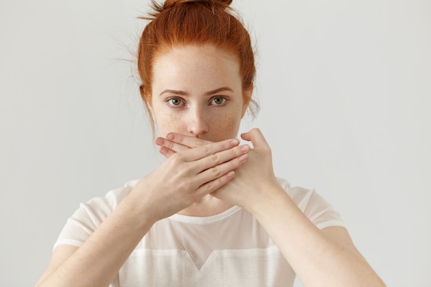 Portret van ernstige jonge gember Europese vrouw die mond behandelt met beide handen die een geheim houden. Sproeterige roodharige vrouw in blouse wil geen geruchten of vertrouwelijke informatie verspreiden