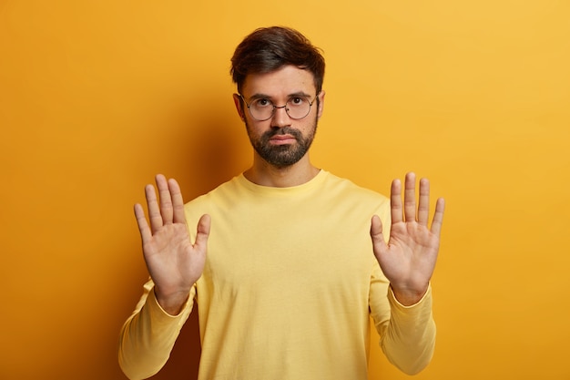 Portret van ernstige ongeschoren man maakt stopsymbool, toont beperking, weigering of afwijzing, vraagt hem niet lastig te vallen draagt een ronde bril en trui geïsoleerd op gele muur heeft waarschuwingsuitdrukking