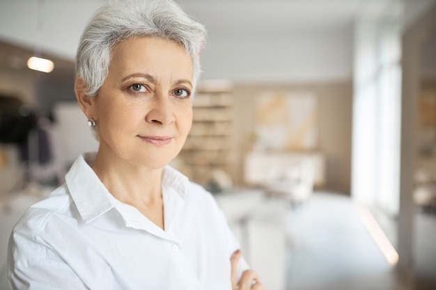 Portret van ernstige zelfverzekerde vrouw van middelbare leeftijd met grijs kort haar, groene ogen, rimpels en charmante glimlach poseren binnenshuis met gevouwen armen