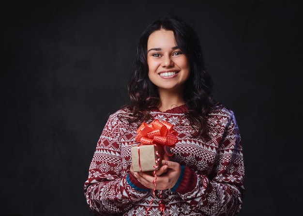 Portret van feestelijk lachende brunette vrouw met lang krullend haar, gekleed in een rode trui houdt kerstcadeaus.