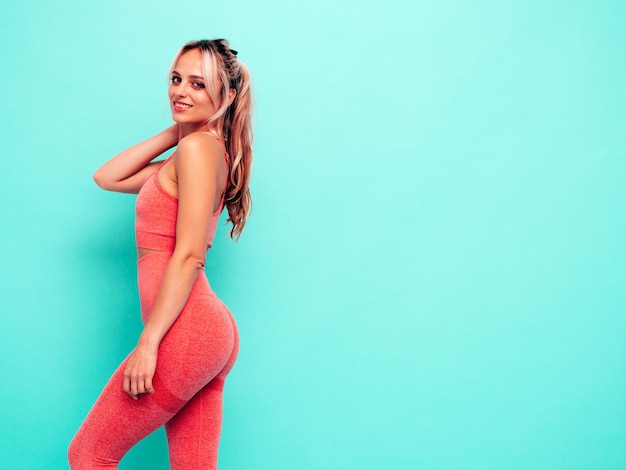 Portret van fitness lachende vrouw in roze sportkleding jonge mooie model met perfect lichaam vrouwelijke poseren in de buurt van blauwe muur in studio vrolijk en gelukkig uitrekken voor de opleiding