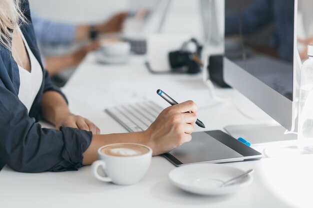 Portret van freelance webdesigner koffie drinken op de werkplek en stylus vasthouden. Licht gebruinde dame in zwart overhemd met behulp van tablet in haar kantoor, achter computer zit.