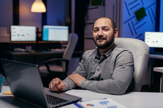 Portret van freelancer die naar camera kijkt die aan bureau zit met grafieken. Slimme zakenman zit op zijn werkplek in de loop van de late nachturen zijn werk te doen.