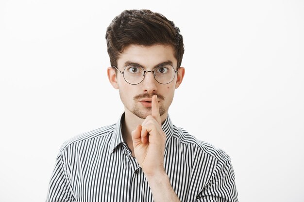 Portret van gefocuste serieuze nerdy kerel in ronde bril, shh zegt terwijl hij een zwijggebaar maakt met wijsvinger voor de mond, nerveuze vriend voelt geheim zal vertellen