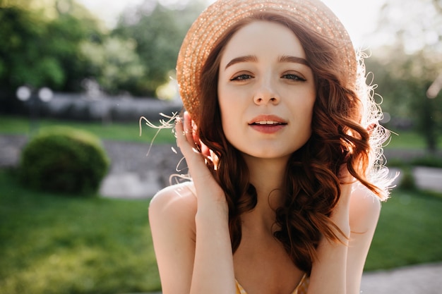 Portret van geïnspireerde gember vrouw in zomer hoed poseren op aard. Gelukkig wit vrouwelijk model met rood haar dat in zonnige dag glimlacht.