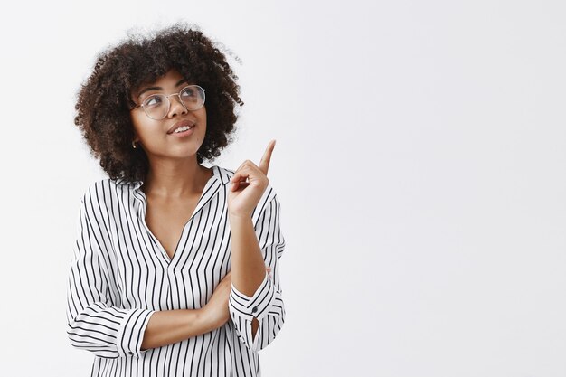 Portret van geïntrigeerde slimme en stijlvolle vrouw in gestreepte blouse en bril kijken en wijzend naar de rechterbovenhoek met nieuwsgierige geïnteresseerde uitdrukking