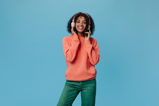 Portret van gekrulde vrouw in oranje outfit geniet van favoriete liedje in koptelefoon Vrij optimistisch meisje in groene denim broek luisteren naar muziek op blauwe achtergrond