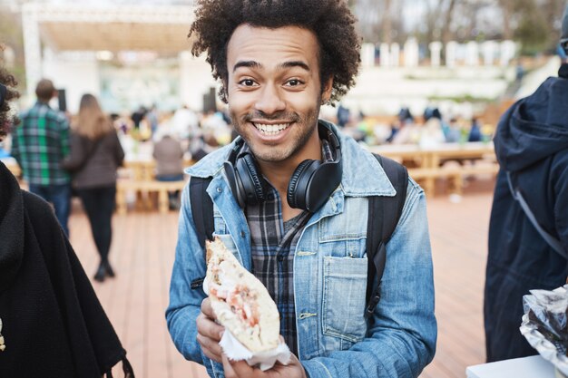 Portret van gelukkig blij Afro-Amerikaanse blogger sandwich houden en glimlachen naar de camera, opgewonden om het te proeven, wandelen over voedselfestival in lokaal park.