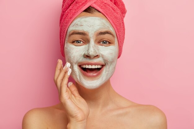 Portret van gelukkig dolblij vrouwelijk model close-up met cosmetische gezichtsmasker aangebracht over haar gezicht, schoonheidsbehandelingen heeft, draagt een handdoek op het hoofd gewikkeld, heeft een gezonde, opgefriste look. Vernieuwing en therapie