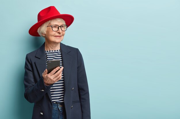 Portret van gelukkig gerimpelde vrouw kijkt opzij, wacht op telefoontje, houdt moderne mobiele telefoons vast, leert hoe moderne technologie te gebruiken, draagt rode hoofddeksels, jas, modellen over blauwe muur, vrije ruimte voor informatie