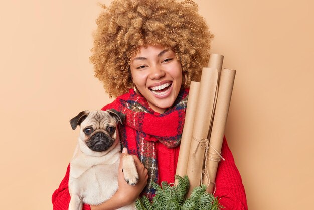 Portret van gelukkig krullend haired vrouw huisdier eigenaar poses met rashond en nieuwjaar decoraties glimlacht ladfully heeft positieve feestelijke stemming gekleed in winterkleren geïsoleerd over bruine achtergrond