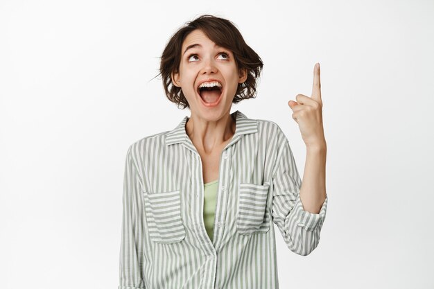 Portret van gelukkig lachende brunette vrouw, kijken en wijzende vinger omhoog staande op wit.