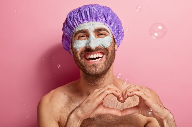 Portret van gelukkig man met brede glimlach, geniet van wassen en schoonheidsbehandelingen, heeft kleimasker op gezicht, staat topless