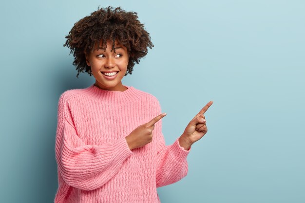 Portret van gelukkig opgetogen vrouw wijst in de goede richting op kopie ruimte, kijkt vrolijk, adverteert met mooi product, draagt een oversized roze trui, geïsoleerd op blauwe muur. Bekijk daar een coole advertentie